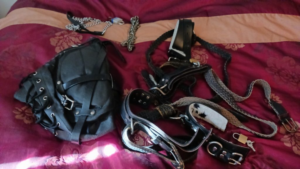 Leather blackout hood, plus a selection of belts, cuffs, and BDSM chains laid out on the bed.