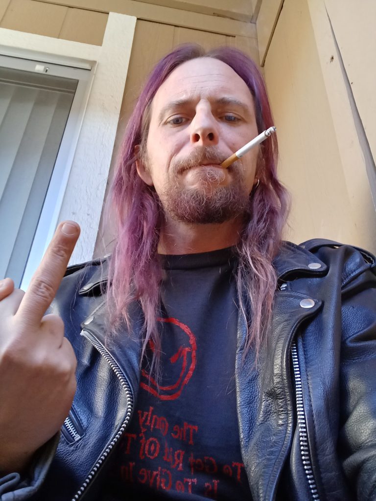 Purple haired nonbinary person with a beard and black leather jacket flips off the camera while wearing a black leather jacket and smoking a cigarette.