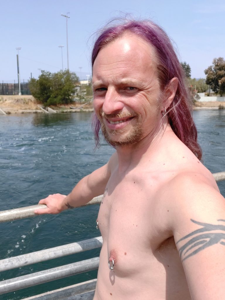 Shirtless at the riverside with purple hair and tattoos.