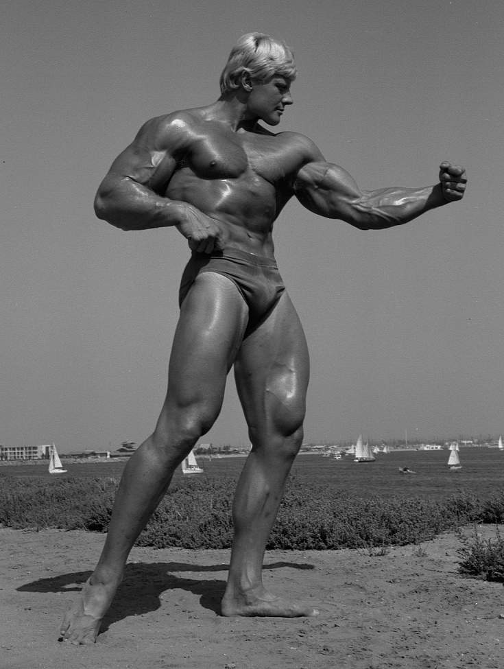 Bodybuilder Dave Draper looks huge against tiny sailboats in the far distance.