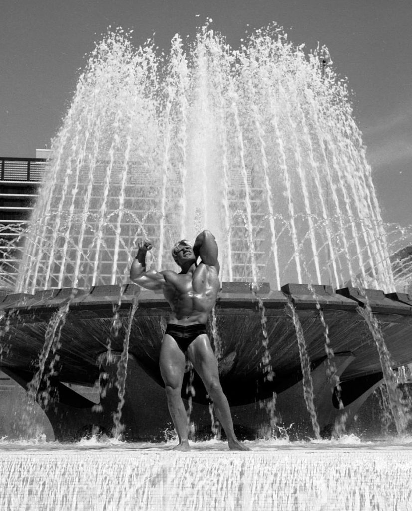 Blonde 60s bodybuilder Dave Draper poses in a fountain.