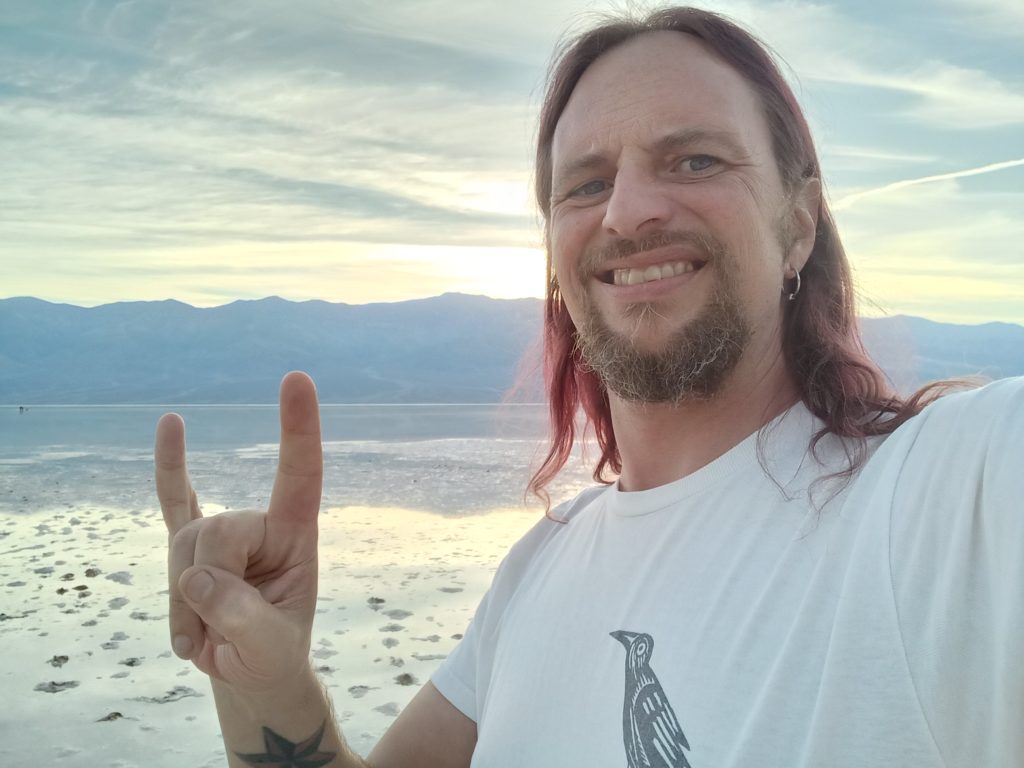 Jack Drago, wearing a T-shirt with a crow on it throws horns in to the camera with a sunset over Lake Manley in the background.