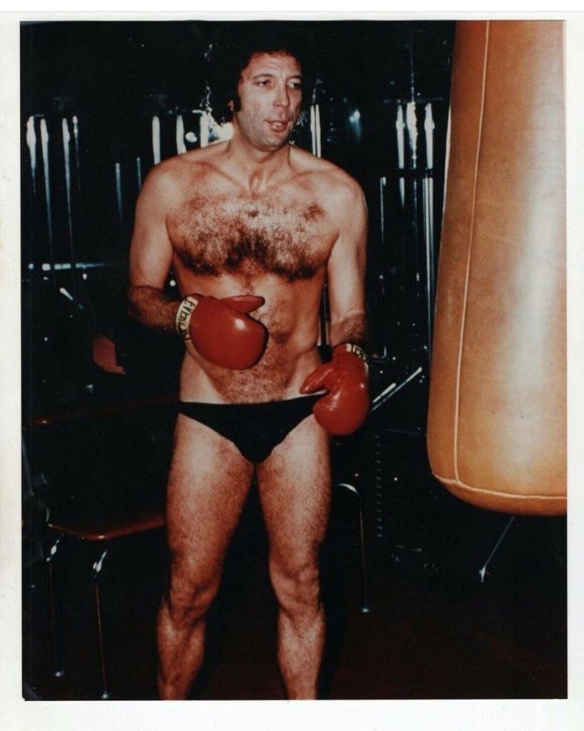 Color photo of a very hairy Tom Jones posing in a skimpy black posing strap in his boxing gloves.