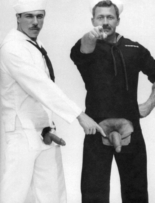 A sailor in a white sailor suit points to the exposed penis of a sailor in a black sailor suit. 