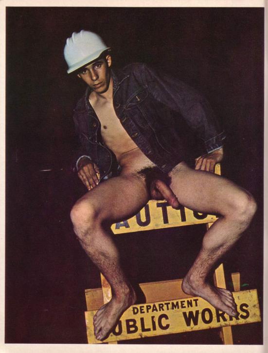 A late 1960s construction worker in a hard hat, jean jacket, and no pants or boots poses on a yellow mid-60s road sign that says "CAUTION: Department of Public Works" with his large, hairy,  circumsized penis exposed and his bare feet partially obscuring the text.