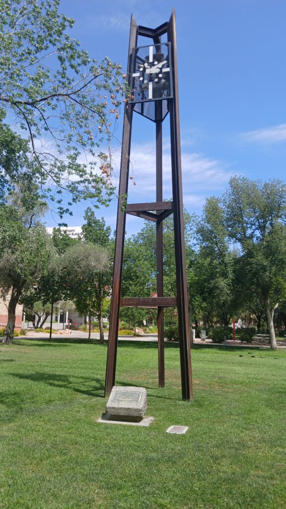 Industrial style clock tower sitting in a grass field.  Analog clock with no numbers just tick marks at 3-6-9-12 reads 10:10. Plaque explains this is the University of Nevada at Las Vegas clock tower installed in 1972