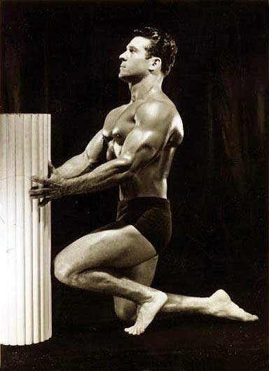 Black and white photo of Jack LaLanne kneeling in black shorts by a white column against a black backdrop.