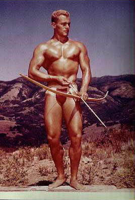 Color photograph of Mike Sill with a bow and arrow in nothing but a red posing strap.